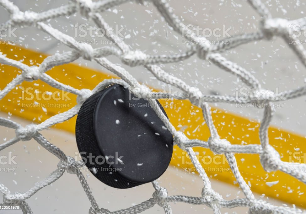 A close-up view of an Ice Hockey puck hitting the back of the goal net as shavings fly by, viewed from the front. Scoring a goal in ice hockey.